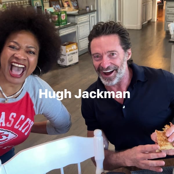 A woman and Hugh Jackman smiling and laughing, with Jackman holding a half-eaten sandwich.