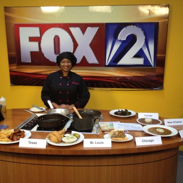 Chef Cathy stands behind a counter with various American dishes and cooking equipment, under a FOX 2 sign.